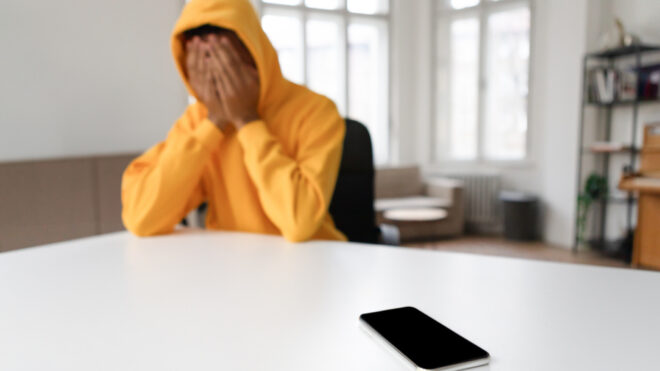A teen boy in distress sits with their head in hands, a smartphone lying on a table
